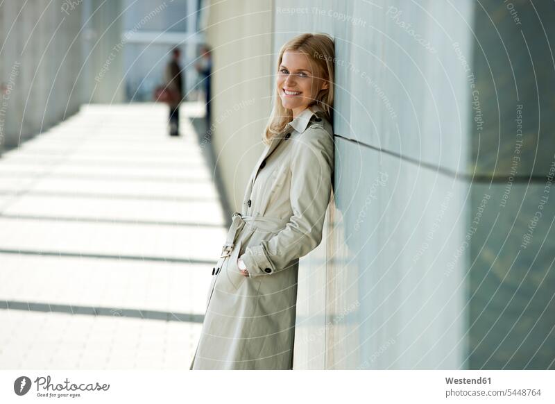 Portrait of smiling blond woman wearing trench coat leaning against facade portrait portraits females women Adults grown-ups grownups adult people persons