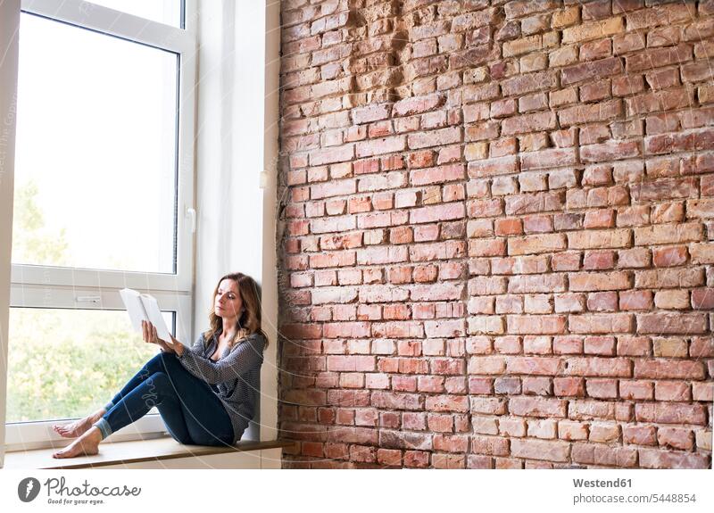 Woman sitting at home on the window sill, reading a book books Seated woman females women windowsill sills window sills Window Cill windowsills Adults grown-ups