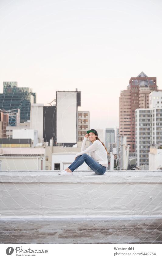 Young woman sitting on balustrade of a rooftop terrace females women parapet happiness happy roof terrace deck carefree smiling smile Adults grown-ups grownups