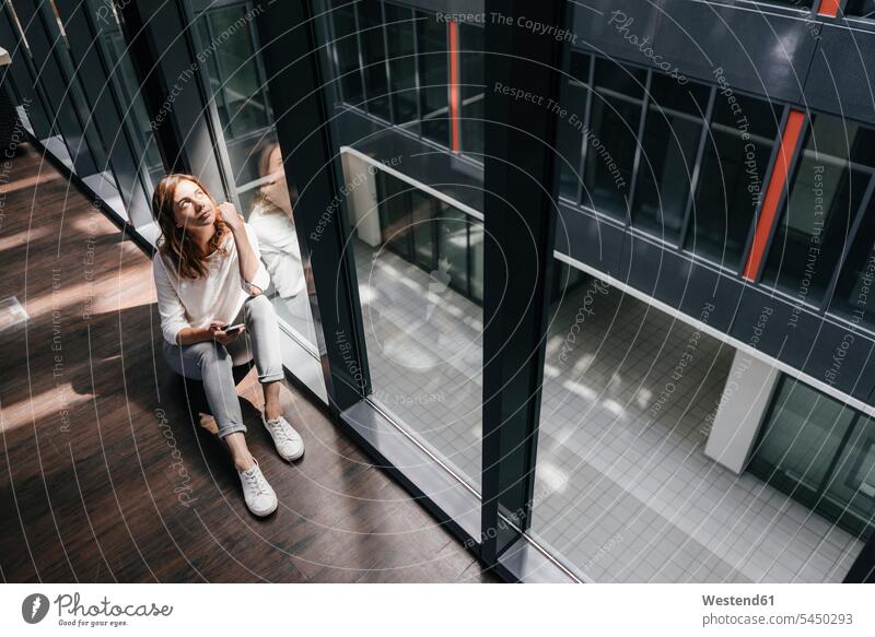 Businesswoman with smartphone, sitting on floor, daydreaming office offices office room office rooms sitting on ground Sitting On The Floor Sitting On Floor