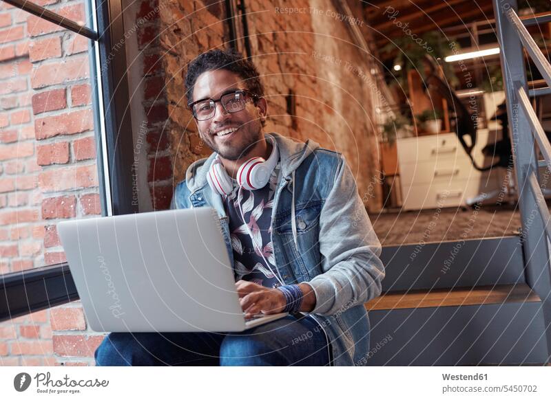 Young man with earphones sitting on office stairs, using laptop young man young men startup young business start up startup company startups young company