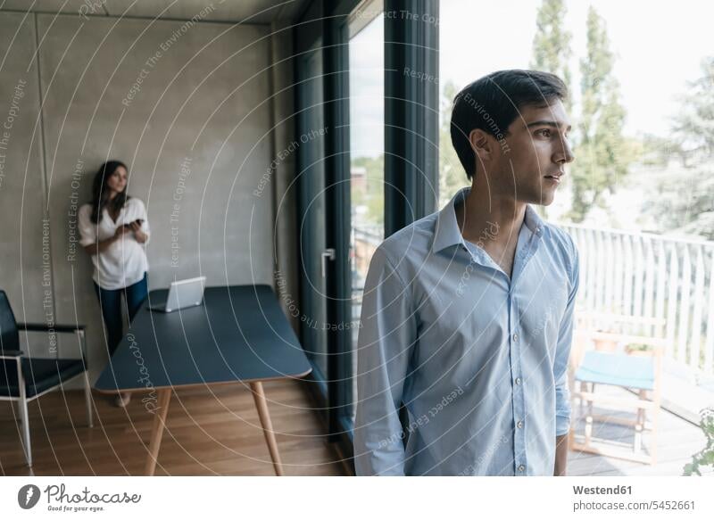 Young man looking out of window with woman in background standing men males windows Adults grown-ups grownups adult people persons human being humans