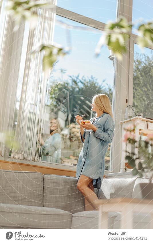 Woman at home standing on couch eating fruit salad looking out of window living room living rooms livingroom Fruit salad settee sofa sofas couches settees woman