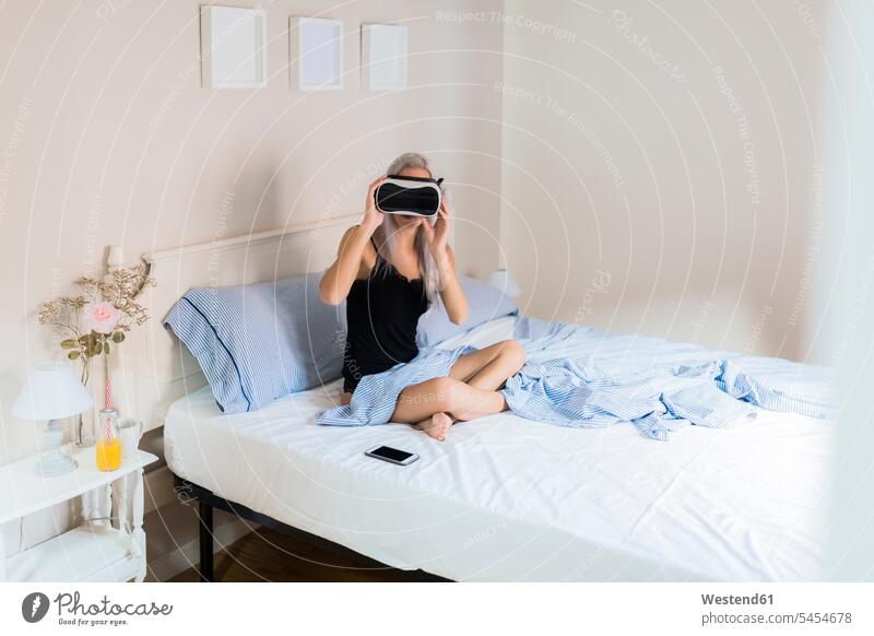 Young woman sitting in bed wearing VR glasses virtual reality females women specs Eye Glasses spectacles Eyeglasses Seated beds Adults grown-ups grownups adult