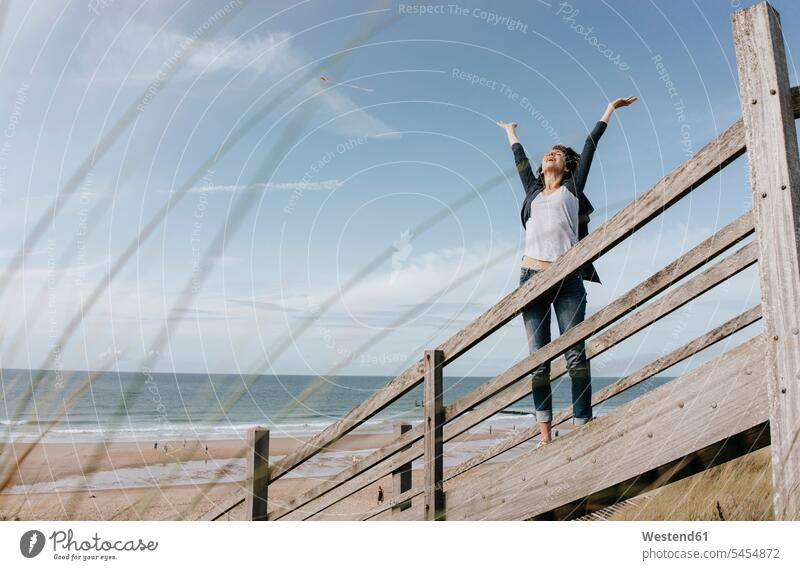 Happy woman standing on boardwalk at the beach with raised arms females women beaches sand dune sand dunes Adults grown-ups grownups adult people persons