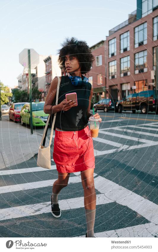 Young woman with headphones and smart phone crossong street in Brooklyn, carying take away drink walking going mobile phone mobiles mobile phones Cellphone