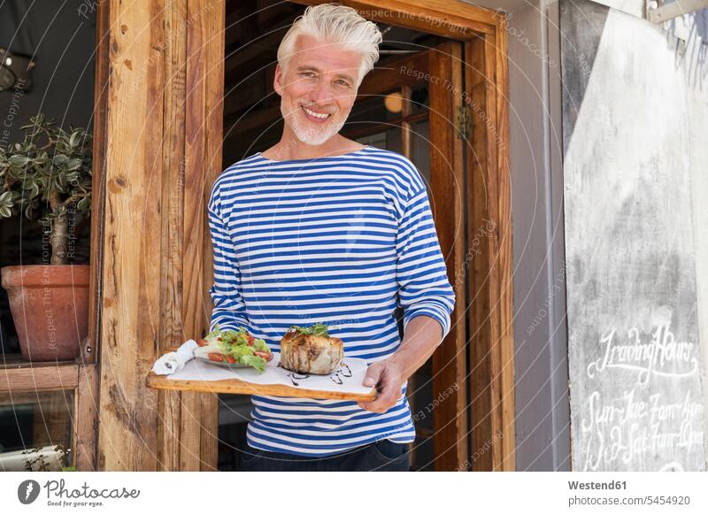 Mature man standing in front of his restaurant, serving a dish door doors owner owners men males restaurants serve happiness happy smiling smile people persons