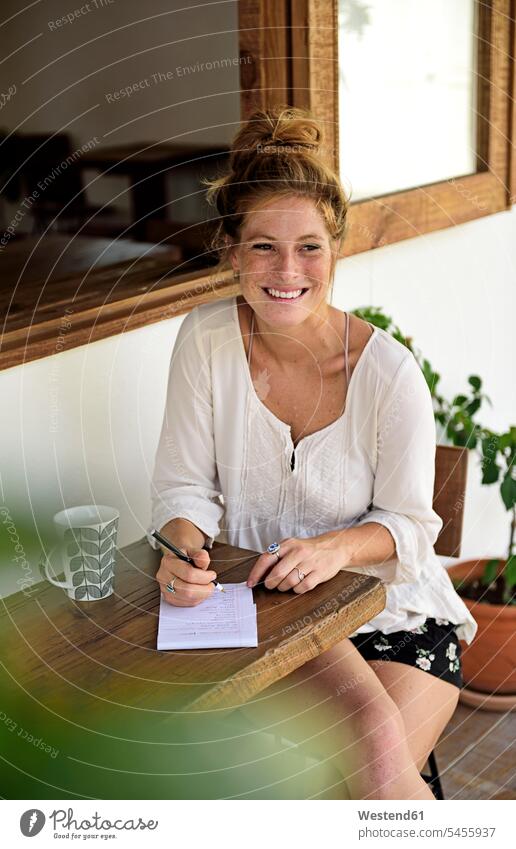 Portrait of laughing young woman with cup of coffee sitting on veranda of a coffee shop writing portrait portraits females women Adults grown-ups grownups adult