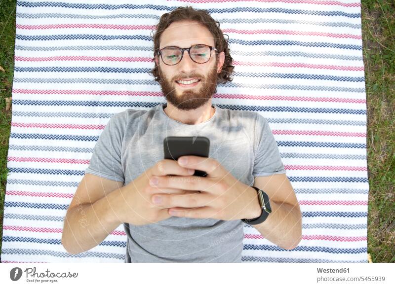 Portrait of smiling man lying on blanket in a park using cell phone, top view portrait portraits men males Smartphone iPhone Smartphones Adults grown-ups