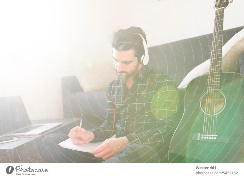 Young man at home with guitar wearing headphones and taking notes guitars couch settee sofa sofas couches settees men males laptop Laptop Computers laptops