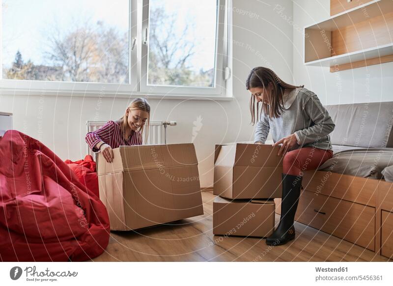 Two young women unpacking cardboard boxes in a room female friends rooms domestic room domestic rooms Cardboard Carton carton Cardboards cartons woman females