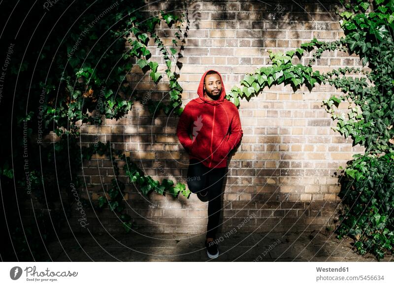Young man wearing red hoodie leaning against a wall brick wall men males standing hooded jacket Hooded Jackets brick walls Adults grown-ups grownups adult