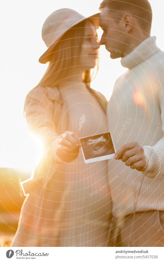 Unrecognised of a pregnant woman with her husband are holding an ultrasound scan photo of the unborn child outdoors on nature in sunlights. Happy motherhood and parenthood concept