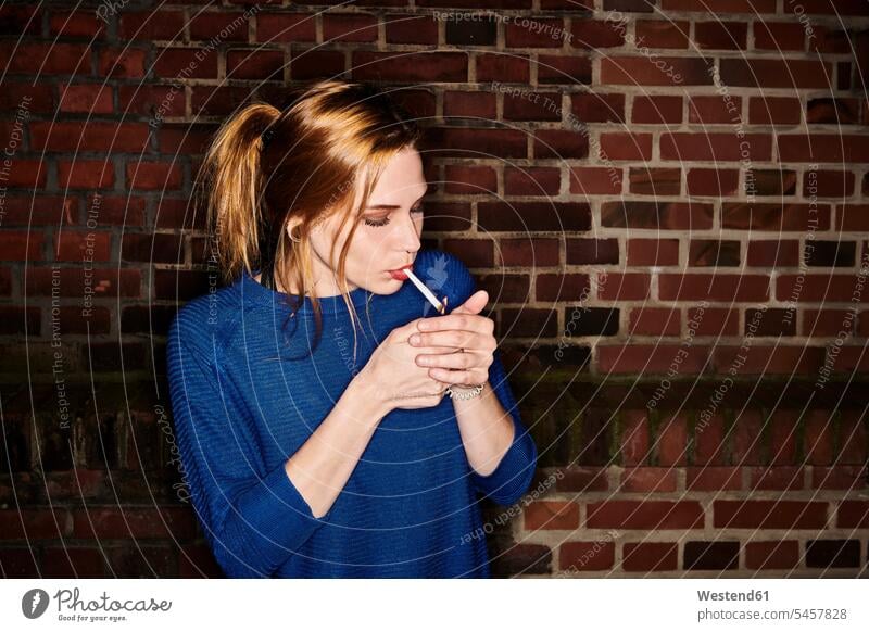 Young woman lighting cigarette against brick wall at night color image colour image outdoors location shots outdoor shot outdoor shots Germany leisure activity