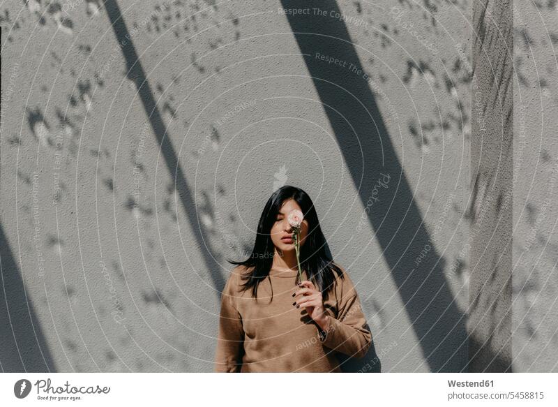 Woman holding flower in front of eye while standing against concrete wall on sunny day color image colour image leisure activity leisure activities free time