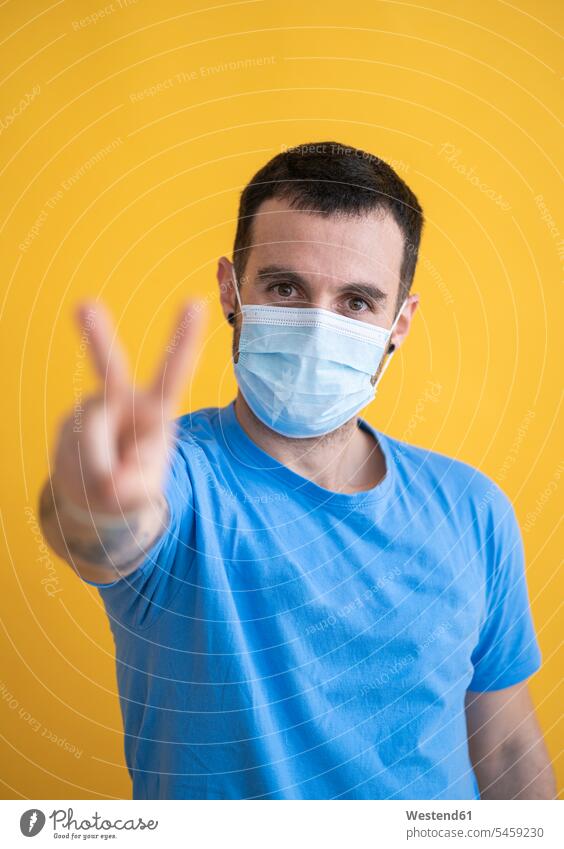 Close-up of man wearing mask showing peace sign while standing against yellow background color image colour image studio shot studio photograph