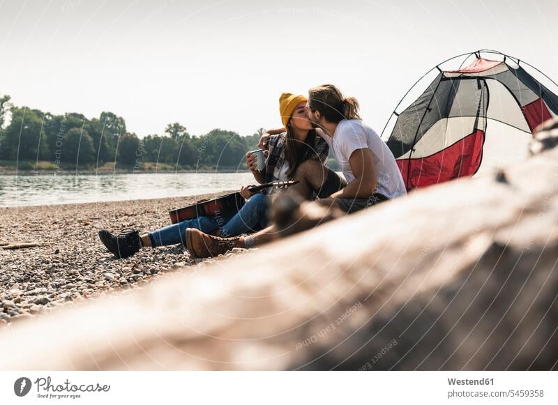 Young couple with guitar kissing at a tent at the riverside tents twosomes partnership couples riverbank sitting Seated kisses guitars people persons