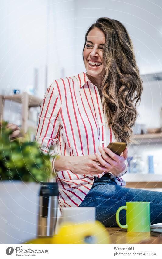 Happy woman with cell phone sitting on table Table Tables females women happiness happy Seated mobile phone mobiles mobile phones Cellphone cell phones Adults