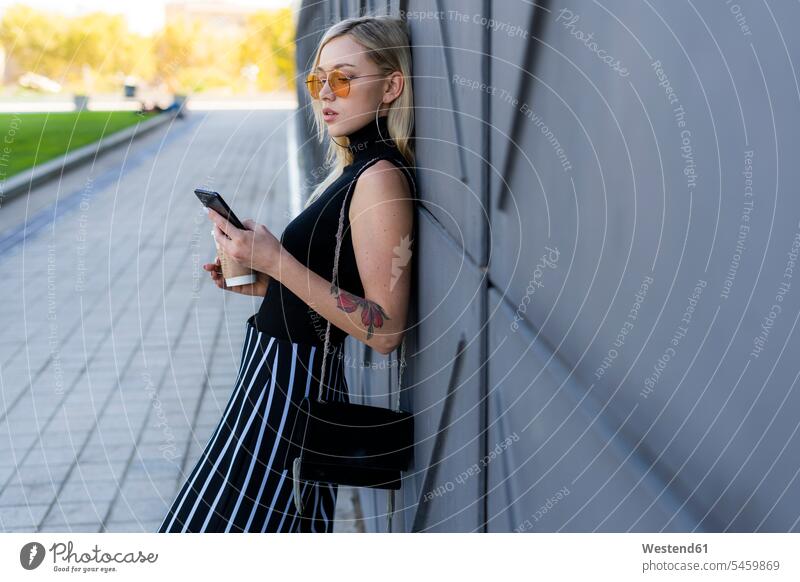 Fashionable young woman leaning against wall looking at smartphone Coffee to Go takeaway coffee technology technologies Technological Connection connected