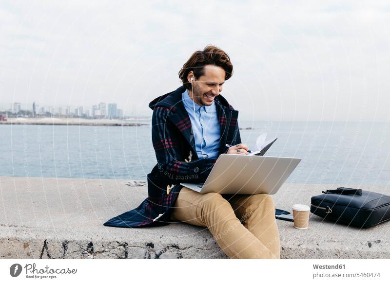 Spain, Barcelona, smiling man sitting at the sea working with laptop and notebook Seated men males notebooks ocean smile At Work Laptop Computers laptops