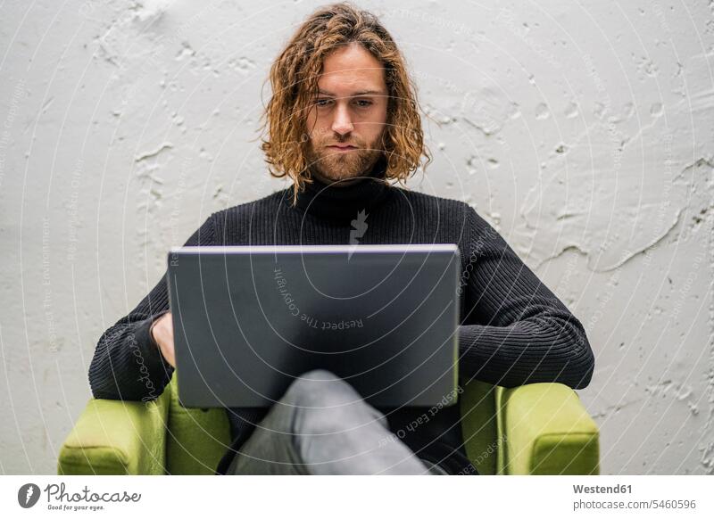Bearded young man using laptop while sitting on armchair against wall at home color image colour image Spain leisure activity leisure activities free time