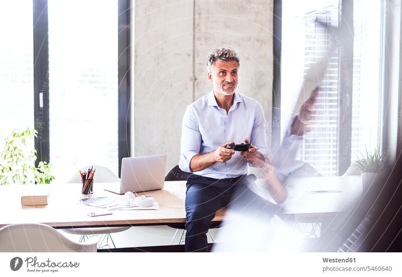 Smiling mature businessman sitting on desk in office using controller offices office room office rooms smiling smile portrait portraits Businessman Business man