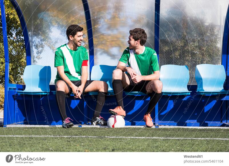 Two happy football players sitting on bench at football field talking happiness footballers soccer players speaking Seated football ground soccer pitch