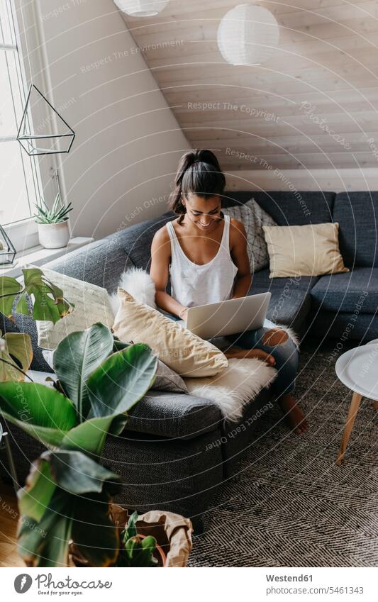 Smiling young woman sitting on the couch at home using laptop use females women Seated smiling smile Laptop Computers laptops notebook settee sofa sofas couches