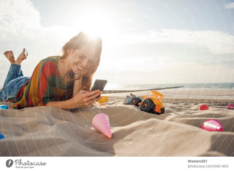 Redheaded woman lying on the beach with beach toys, using smartphone beaches Smartphone iPhone Smartphones females women playing redheaded red hair red hairs