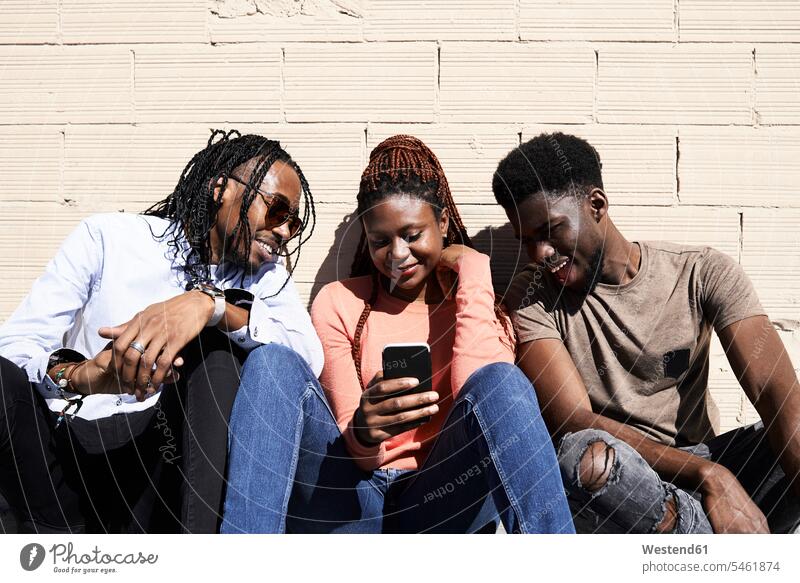 Three friends sitting together and watching a video on a smartphone outdoors happiness happy looking looking at video film mobile phone mobiles mobile phones
