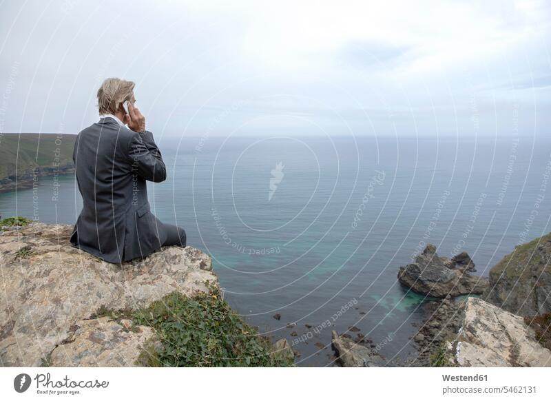 UK, Cornwall, Gwithian, businessman sitting at the coast talking on cell phone coastline shoreline on the phone call telephoning On The Telephone calling Seated