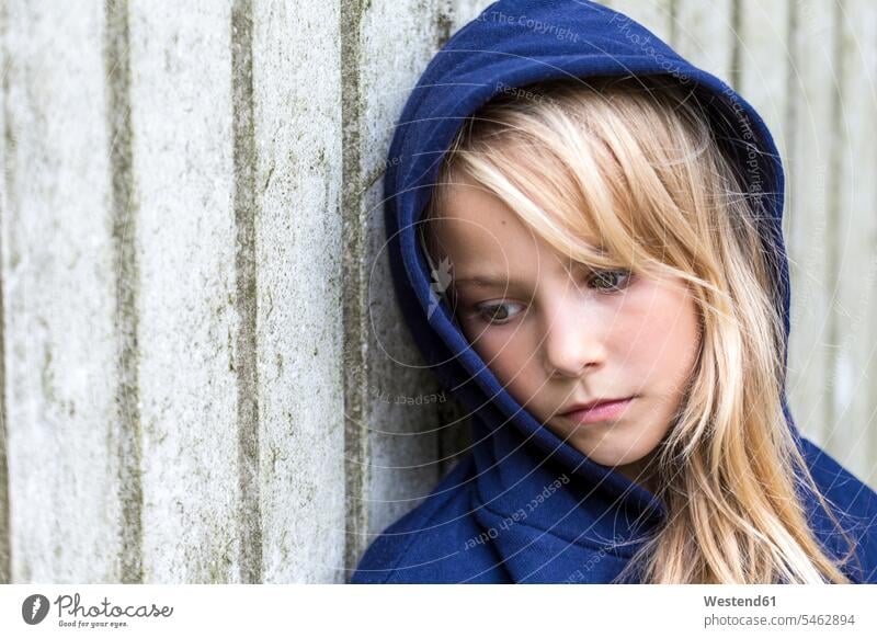 Portrait of sad blond girl wearing blue hooded jacket leaning against wooden wall females girls portrait portraits Hooded Jackets blond hair blonde hair child