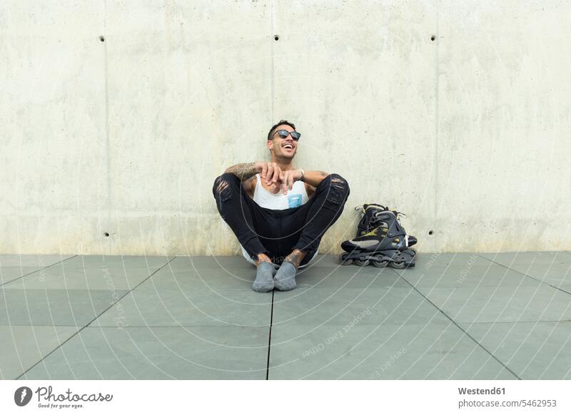Tattooed young man with roller skates sitting on ground having fun inline-skater inline-skaters Roller-Skate tattoo tattoos land Fun funny men males Seated