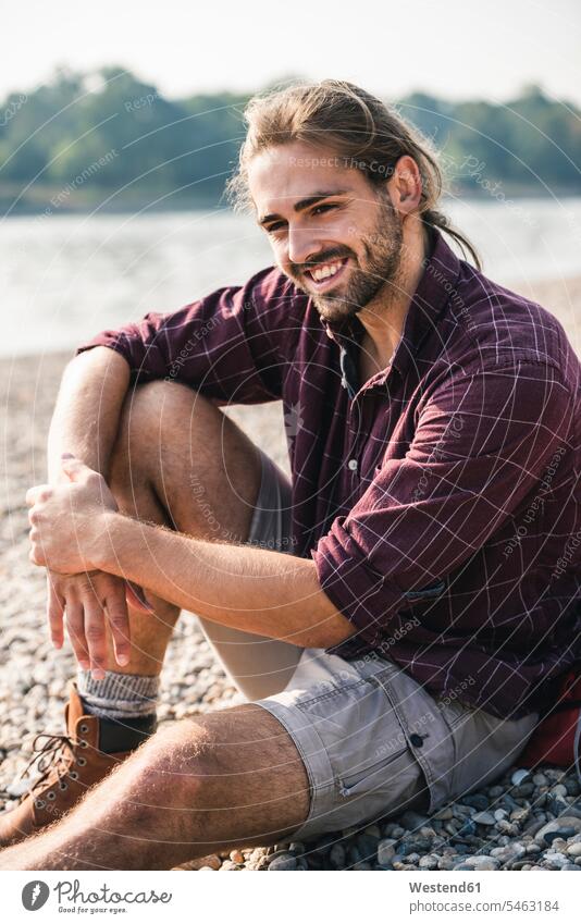 Smiling young man sitting at the riverside River Rivers smiling smile Seated men males riverbank water waters body of water Adults grown-ups grownups adult