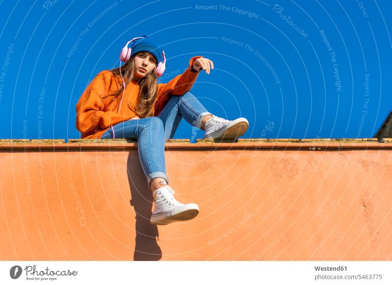 Portrait of teenage girl sitting on wall against sky listening music with headphones jumper sweater Sweaters headset hear Seated colour colours free time