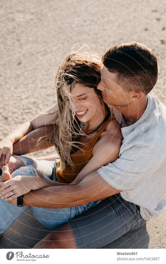 Happy affectionate young couple sitting on the beach touristic tourists cuddle snuggle snuggling smile Seated embrace Embracement hug hugging summer time