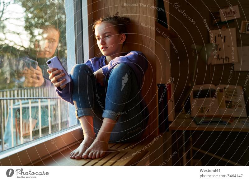 Girl sitting barefoot on window sill in the evening looking at smartphone Tables desks telecommunication phones telephone telephones cell phone cell phones