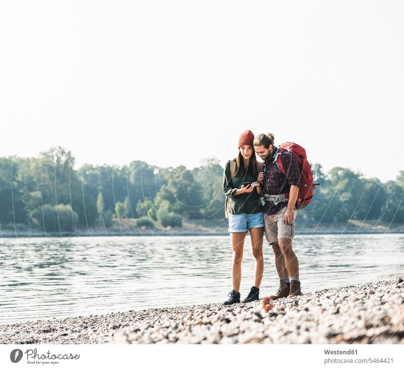 Young couple with backpacks at the riverside checking cell phone rucksacks back-packs River Rivers twosomes partnership couples riverbank mobile phone mobiles