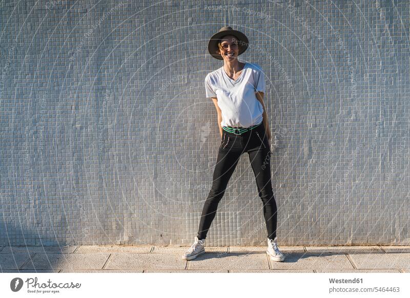 Laughing woman wearing hat standing in front of tiled wall at sunlight hats females women laughing Laughter Sunlit walls Adults grown-ups grownups adult people