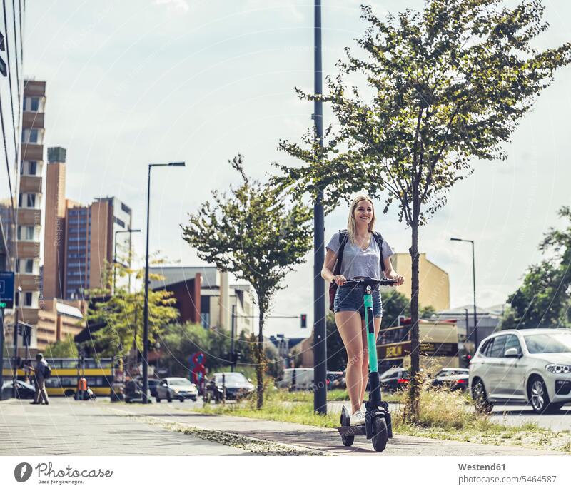 Smiling young woman riding E-Scooter on bicycle lane, Berlin, Germany back-pack back-packs backpacks rucksack rucksacks motor vehicles road vehicle