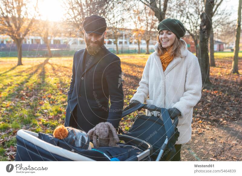 Happy couple with little son in a pushchair at park coat coats jackets caps hat hats baby carriage baby carriages prams stroller strollers smile delight