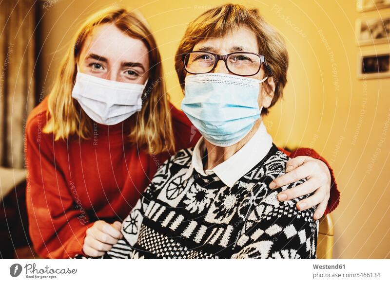 Granddaughter sitting with arm around on grandmother wearing face mask at home during Covid-19 color image colour image indoors indoor shot indoor shots