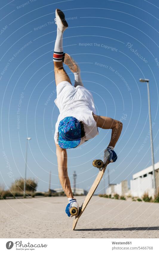 Back view of man in stylish sportive outfit standing on skateboard upside down against blue sky Trick skateboarding rear view back view view from the back