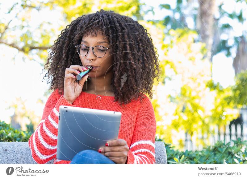 Afro woman smoking electronic cigarette while using digital tablet in public park color image colour image outdoors location shots outdoor shot outdoor shots