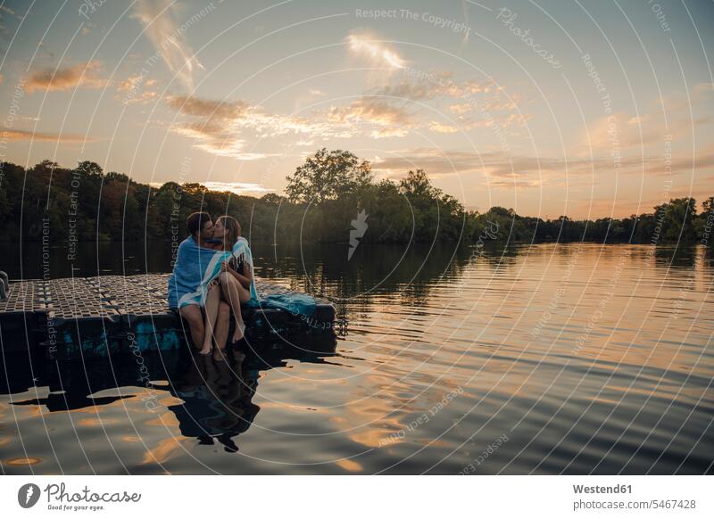 Young couple sitting on bathing platform, kissing, wrapped in towel relax relaxing Seated embrace Embracement hug hugging in the evening summer time summertime