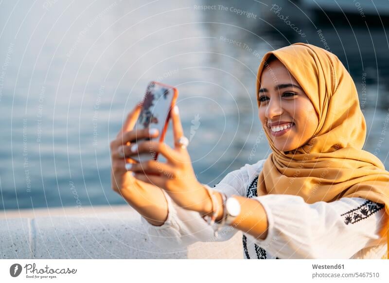 Young muslim woman wearing yellow hijab and using smartphone and taking a selfie human human being human beings humans person persons North African 1