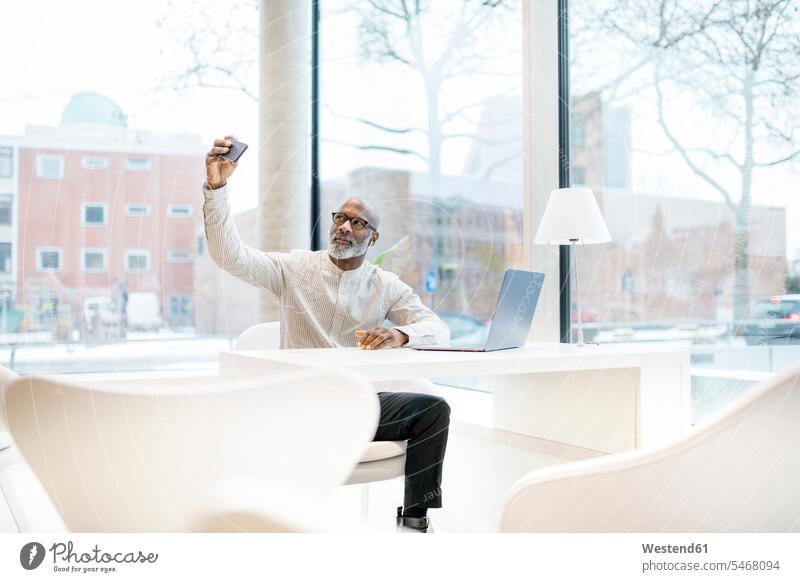 Portrait of mature man with laptop sitting at desk taking selfie with smartphone Businessman Business man Businessmen Business men portrait portraits Smartphone