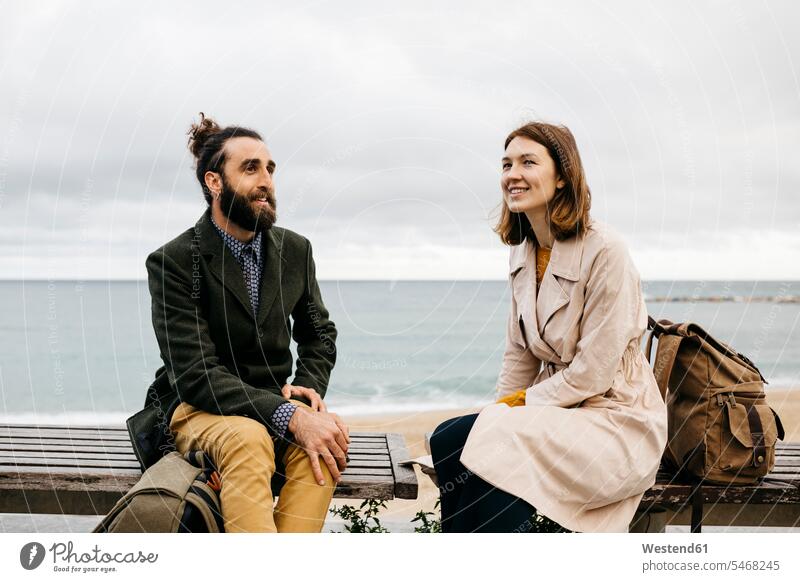 Smiling couple sitting on a bench at beach promenade talking speaking sea front boardwalk break twosomes partnership couples Seated smiling smile benches