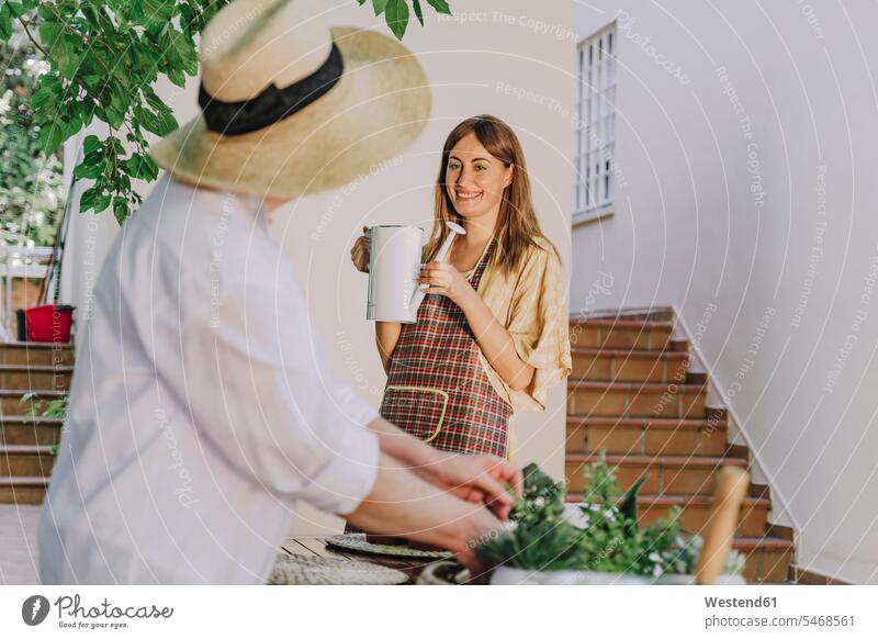 Senior woman wearing hat looking at daughter holding watering can against house in yard color image colour image Spain leisure activity leisure activities