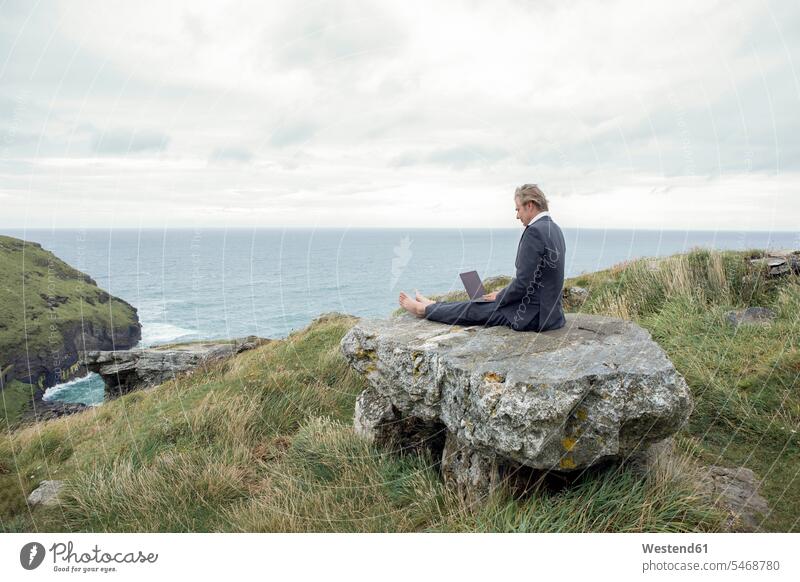 UK, Cornwall, Tintagel, businessman sitting on rock at the coast using laptop Businessman Business man Businessmen Business men Laptop Computers laptops